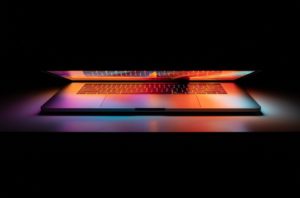 En nästan ihopfälld silverfärgad laptop mot svart bakgrund. Runt den återges skärmens ljus i orange och blå nyanser
